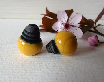 May include: Two ceramic beads, one yellow with black stripes and one yellow with a black top, are on a white surface. The beads are round and have a glossy finish.