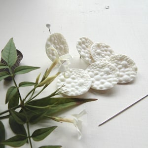 Peut inclure: Boutons en céramique blanche à surface texturée, disposés en grappe sur un fond blanc. On voit également une brindille de fleurs de jasmin blanches et de feuilles vertes.