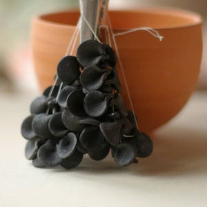 Op de afbeelding: Een cluster van zwarte keramische bloemvormige bedels die aan een terracotta kom hangen. De bedels zijn aan elkaar geregen met wit garen.