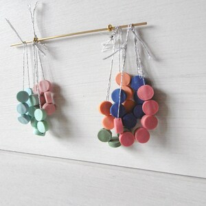 Puede incluir: Un conjunto de cuentas redondas de colores enhebradas en finos cordones blancos, colgando de una varilla dorada. Las cuentas son en tonos de azul, verde, rosa y naranja.