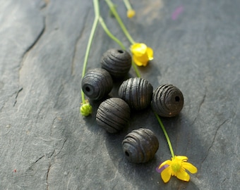 May include: Six dark grey wooden beads with grooved lines, alongside yellow flowers and green stems on a dark grey stone surface. The spherical beads have a matte finish, suggesting a natural, handcrafted aesthetic. Each bead is approximately 1.5 cm in diameter.
