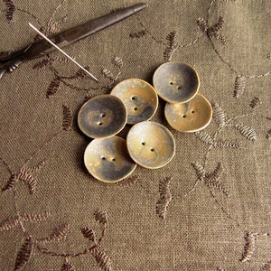 May include: Six ceramic buttons in shades of brown and gold. The buttons are round and have a slightly raised center. They are arranged in a semi-circle on a brown fabric background.