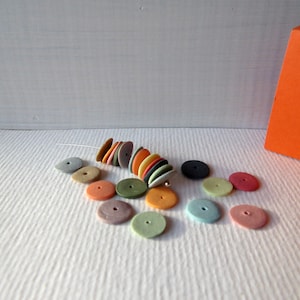 Puede incluir: Una pila de cuentas redondas de cerámica de colores en varios tonos de naranja, amarillo, verde, azul y gris. Las cuentas están dispuestas en espiral y están esparcidas sobre una superficie blanca.