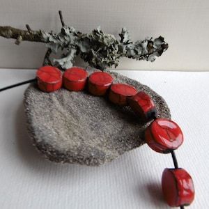 Peut inclure: Un collier de perles en céramique rouge avec un cordon noir. Les perles sont rondes et ont une surface texturée. Le collier est présenté sur un morceau de bois flotté.