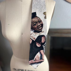Può includere: Una cravatta tessuta con un ritratto. La cravatta presenta uno sfondo chiaro con un motivo ripetuto e un'immagine dettagliata di una persona. La cravatta è esposta su un manichino.