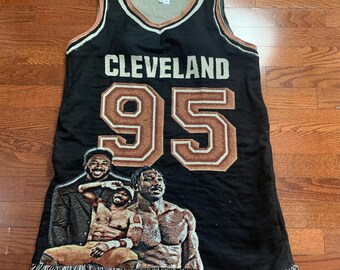 May include: A black sleeveless jersey-style top featuring the text CLEVELAND and the number 95 in brown and white. The garment displays a woven graphic of three individuals and includes a fringed hemline at the bottom edge of the piece.