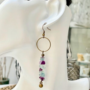 May include: A pair of dangle earrings with a brass hoop and a chain of light blue, purple, and green fluorite stones. The earrings are hanging from a mannequin's ear.