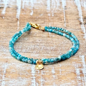 Può includere: Un delicato braccialetto a doppio filo con perline di pietre preziose blu turchese e un ciondolo dorato. Il braccialetto è realizzato con una chiusura dorata ed è perfetto per essere sovrapposto ad altri gioielli.