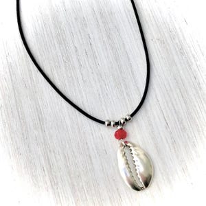 Op de afbeelding: Een zilveren schelp hanger ketting met een rode kraal en zilveren kralen op een zwart koord.