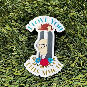 Op de afbeelding: Een witte sticker met een cartoonillustratie van een man die een rode pruik omhoog houdt. De tekst op de sticker luidt "Ik hou van je zo veel".