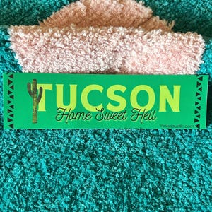 May include: Green and gold sticker with the text "Tucson Home Sweet Hell" and a cactus graphic. The sticker is on a pink and teal fuzzy surface.