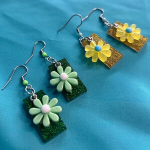 Puede incluir: Un par de pendientes rectangulares de purpurina verde con un dije de flor blanca y un par de pendientes rectangulares de purpurina amarilla con un dije de flor azul y amarilla.