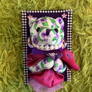 May include: A plush bear with a white body, adorned with purple and green floral patterns. The bear is dressed in a pink dress and a pearl necklace, set against a purple background with a beaded border.