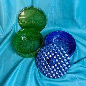 May include: A two-piece, round, decorative box with a green lid and a blue lid. The green lid has a geometric pattern and the blue lid has a diamond pattern. The box is made of resin and has a glittery finish.