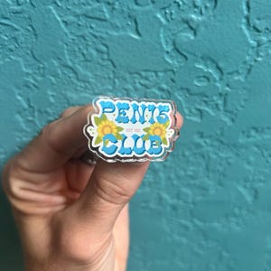 May include: A clear acrylic pin with the words "PENIS CLUB" in blue letters with yellow flowers on either side.