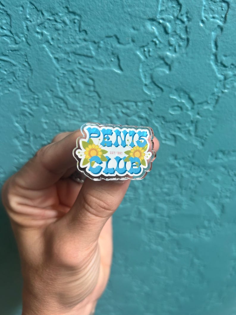 May include: A clear acrylic pin with the words "PENIS CLUB" in blue letters with yellow flowers on either side.