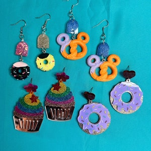 May include: A set of colorful, glittery earrings featuring various food-themed designs, including donuts, pretzels, cupcakes, and a sprinkle-covered treat.