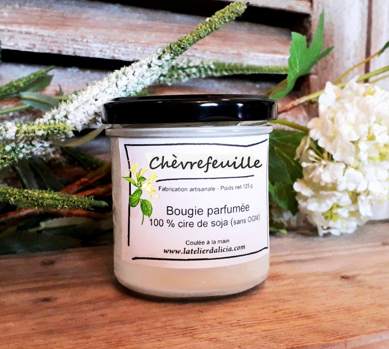 Pu&ograve; includere: Una candela di vetro in un barattolo con un'etichetta bianca che recita "Ch&egrave;vrefeuille" e "Bougie parfum&eacute;e 100% cire de soja (sans OGM)". L'etichetta include anche il testo "Fabrication artisanale - Poids net 125g" e "Coul&eacute;e &agrave; la main" e il sito web "www.latelierdalicia.com".