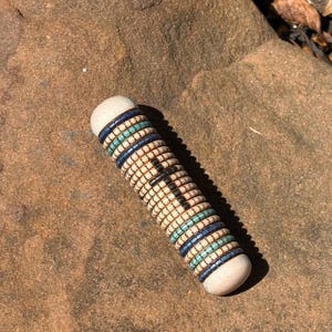 May include: A cylindrical beaded object with light-colored wooden ends. The body is adorned with alternating rows of small, square beads in cream, blue, and turquoise. The beads are arranged in a spiral pattern, creating a textured surface.