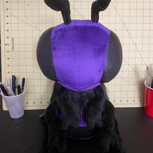 May include: A purple and black plush toy shaped like a bee with large, black antennae. The bee is sitting on a black surface.