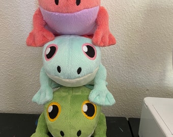 May include: Three plush frog toys stacked on top of each other. The top frog is coral with a lavender mouth, the middle frog is light blue, and the bottom frog is green. Each frog has large eyes.