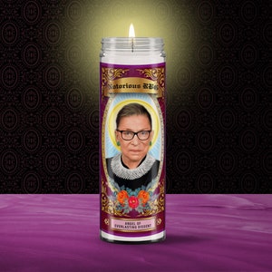 May include: A white candle with a purple label featuring a portrait of Ruth Bader Ginsburg. The label reads "Notorious RBG" and "Angel of Everlasting Dissent".