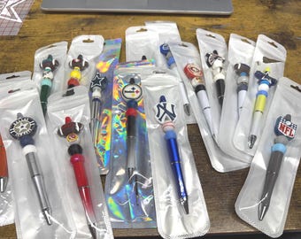 Puede incluir: Una colección de bolígrafos variados, cada uno empaquetado individualmente en fundas de plástico transparente. Los bolígrafos cuentan con cuentas de silicona decorativas y varios logotipos de equipos deportivos, incluidos emblemas de la NFL y MLB, en diversos colores.