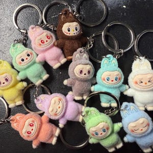 May include: A collection of colorful plush keychains featuring a cartoon character with a smiling face and bunny ears. The keychains are in various colors, including pink, yellow, green, and blue, attached to silver key rings.