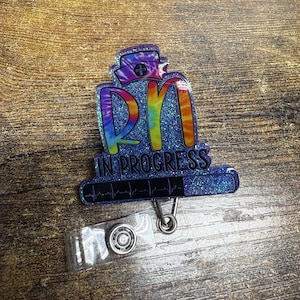 Puede incluir: Un portatarjetas RN azul brillante con un diseño de gorro de enfermera teñido anudado. Las letras "RN" son de colores arcoíris, y el texto "IN PROGRESS" está encima de un gráfico de latidos. Incluye un clip de carrete de plástico transparente.