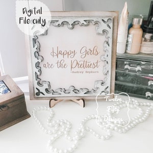 May include: A white framed sign with a decorative border and the quote "Happy girls are the prettiest -Audrey Hepburn" in brown text. The sign is sitting on a wooden stand and there are white pearl beads in the foreground.