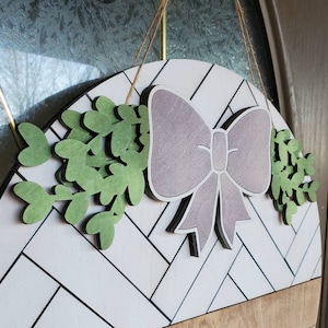 May include: A white and brown wooden door wreath with a chevron pattern. The wreath is decorated with a large purple bow and green leaves.