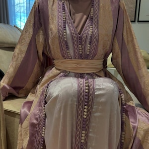 K&ouml;nnte beinhalten: Ein langes, flie&szlig;endes Kleid in den Farbt&ouml;nen Lavendel, Gold und Creme. Das Kleid hat komplizierte Goldmuster, vertikale Lavendelstreifen und einen cremefarbenen G&uuml;rtel. Der Ausschnitt und die Kanten sind mit kleinen, wei&szlig;en Perlen verziert.