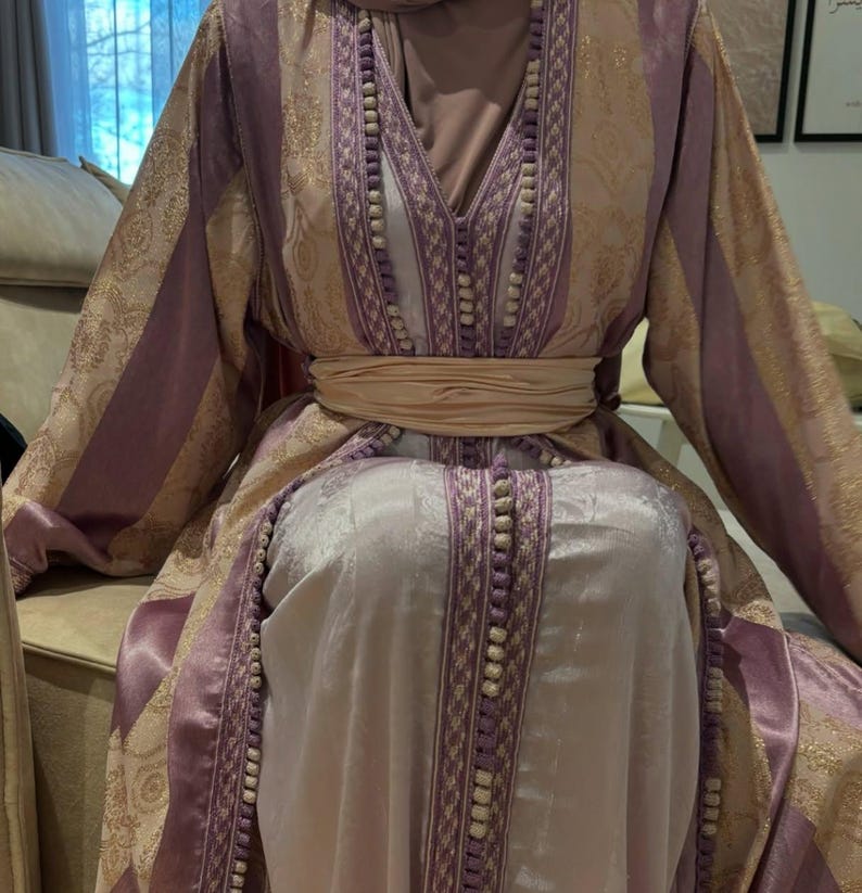 K&ouml;nnte beinhalten: Ein langes, flie&szlig;endes Kleid in den Farbt&ouml;nen Lavendel, Gold und Creme. Das Kleid hat komplizierte Goldmuster, vertikale Lavendelstreifen und einen cremefarbenen G&uuml;rtel. Der Ausschnitt und die Kanten sind mit kleinen, wei&szlig;en Perlen verziert.