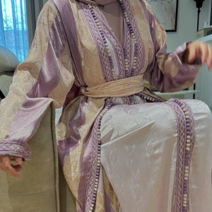 K&ouml;nnte beinhalten: Traditionelles Kleidungsst&uuml;ck in Helllila und Creme mit aufwendiger Stickerei und passender Kopfbedeckung. Das Kleidungsst&uuml;ck hat eine betonte Taille und dekorative Verzierungen. Die &Auml;rmel sind weit und flie&szlig;end, und der Stoff hat einen dezenten Glanz.