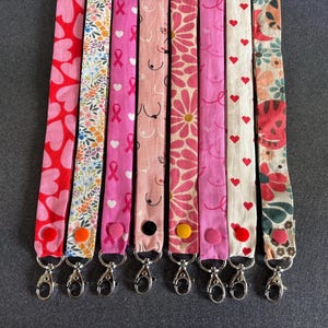 Può includere: Otto cordini in tessuto disposti verticalmente, ognuno con motivi distinti tra cui cuori rossi, stampe floreali, nastri rosa per la consapevolezza del cancro al seno, linee astratte, margherite rosa, rosa tinta unita e cuori rossi su bianco.
