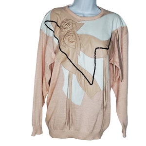 Peut inclure: Un pull rose avec une applique en cuir blanc et beige représentant un motif de rose. L'applique est ornée de franges.