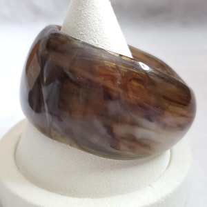 May include: A wide, thick band ring featuring a polished, semi-translucent finish with a marbled pattern of dark brown, tan, and cream swirls. The ring has a smooth, rounded exterior surface and a glossy appearance, showcasing a continuous, fluid design.