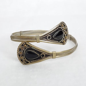 May include: An antique-style arm cuff bracelet with a silver-tone finish. The bracelet features two ornate, teardrop-shaped black stone settings with gold-tone accents. The cuff is designed to wrap around the arm.