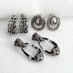 Könnte beinhalten: Eine Sammlung silberfarbener Ohrringe. Die Ohrringe umfassen zwei Paar ovale Clip-Ohrringe mit schwarzem Emaille-Akzent und dekorativem Aufsatz, zwei Paar runde Clip-Ohrringe mit Perlendesign und zwei Paar verzierte, vertikale Ohrringe.