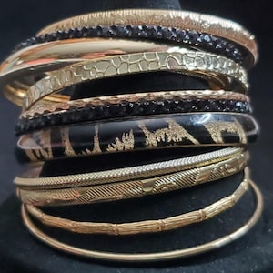 May include: A stack of nine gold-toned and black bangles. The set includes smooth gold bands, textured gold bands, two bands encrusted with small black stones, and one wide black band featuring a gold-coloured glittery pattern of animal silhouettes.
