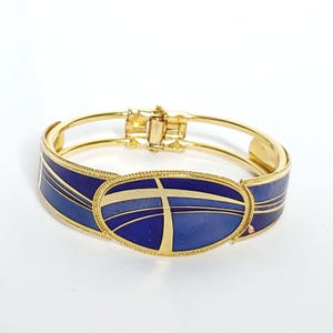 May include: Gold-toned hinged bracelet with a central oval design. The bracelet features a deep blue enamel inlay with gold accents. The oval is bisected by a gold cross design. The bracelet has a secure clasp.