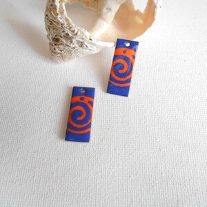 May include: Two rectangular blue earrings with orange spiral designs. Each earring has a small hole at the top. The earrings are displayed on a white surface with a seashell in the background.