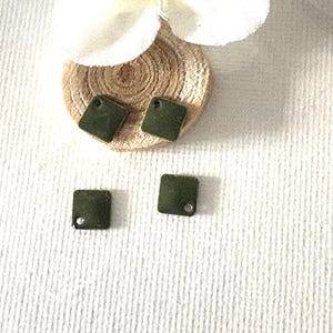 Peut inclure: Trois boucles d'oreilles carrées vert olive avec de petits trous pour les accrocher. Deux boucles d'oreilles sont placées sur un petit disque rond en bois brun clair. Une fleur blanche est placée au-dessus du disque en bois. Les boucles d'oreilles sont sur une surface blanche texturée.