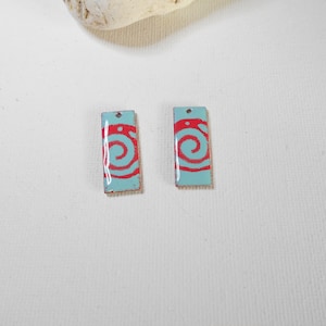May include: Two rectangular enamel charms with a light blue background and a red spiral design.
