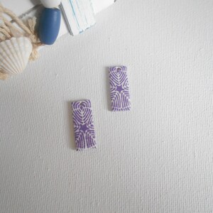 May include: Two purple and white ceramic rectangular tiles with a geometric pattern. The tiles have a hole at the top for hanging.