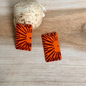 May include: Two rectangular pendants featuring a vibrant orange background with a dark brown sunburst pattern radiating from the side. Each piece has a small hole at the top for attachment, showcasing a flat, smooth finish and a bold graphic design.