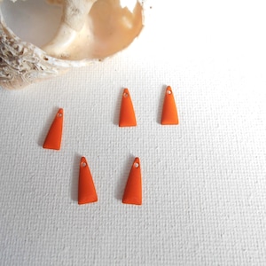 Peut inclure: Cinq éléments de bijouterie orange de forme triangulaire avec un petit trou en haut. Les éléments sont disposés sur une surface blanche, avec un coquillage en arrière-plan. Ces éléments sont probablement utilisés pour la fabrication de bijoux.