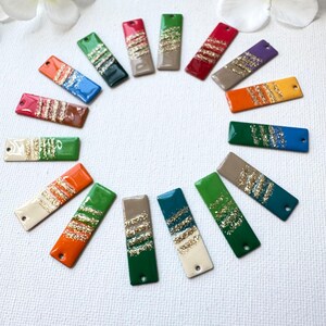 May include: A collection of rectangular pendants in various colors, including red, green, and blue, each with a glittery gold stripe design. The pendants are arranged in a circular pattern.