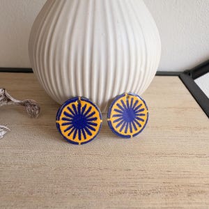 Peut inclure: Deux boucles d'oreilles rondes avec un motif de soleil bleu et jaune. Les boucles d'oreilles sont présentées sur un fond neutre, avec un vase blanc en arrière-plan.