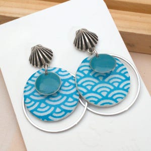 Könnte beinhalten: Ein Paar Ohrringe mit einem muschelförmigen Stecker und einem silbernen Reifen. Die Ohrringe haben ein türkisfarbenes und weißes kreisförmiges Design mit einem kleineren türkisfarbenen Kreis oben. Die Ohrringe befinden sich auf einer weißen Oberfläche.